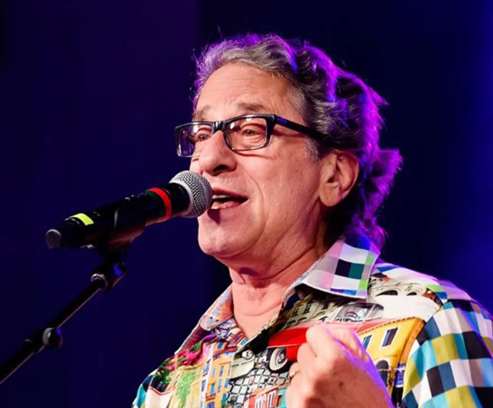 A person with glasses is speaking into a microphone against a purple background wearing a colorful checked shirt