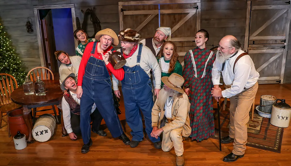 A group of actors in a rustic wooden set are posing joyfully in various costumes that suggest a play set in a bygone era with some in festive attire and others portraying more comical characters