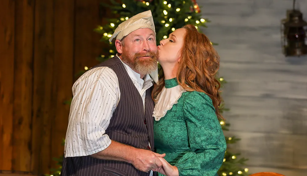 A man and a woman dressed in historical costumes appear to share an affectionate moment onstage with the woman kissing the man on the cheek