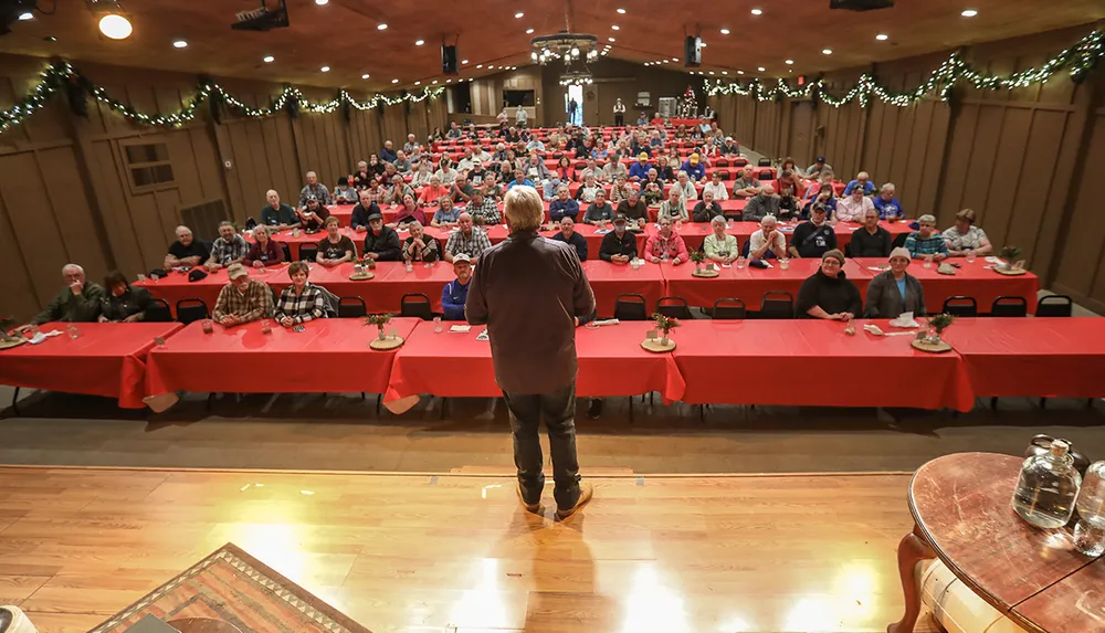 A person is standing on a stage speaking to a seated audience arranged at tables with red tablecloths in a well-lit banquet hall decorated with string lights