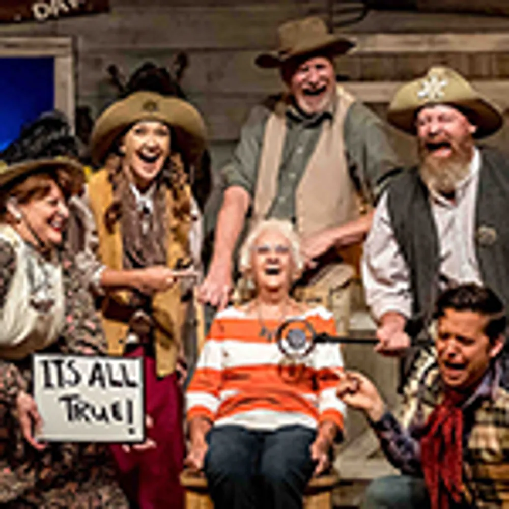 A group of six people dressed in Western-themed costumes are playfully gathered around an older woman who is sitting and smiling with one person holding a sign that says ITS ALL TRUE