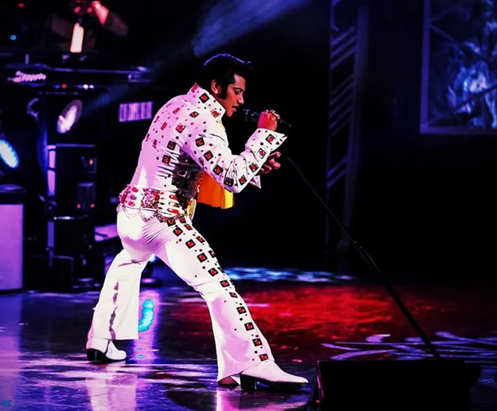 A performer dressed in an Elvis Presley-inspired jumpsuit is singing on stage into a microphone