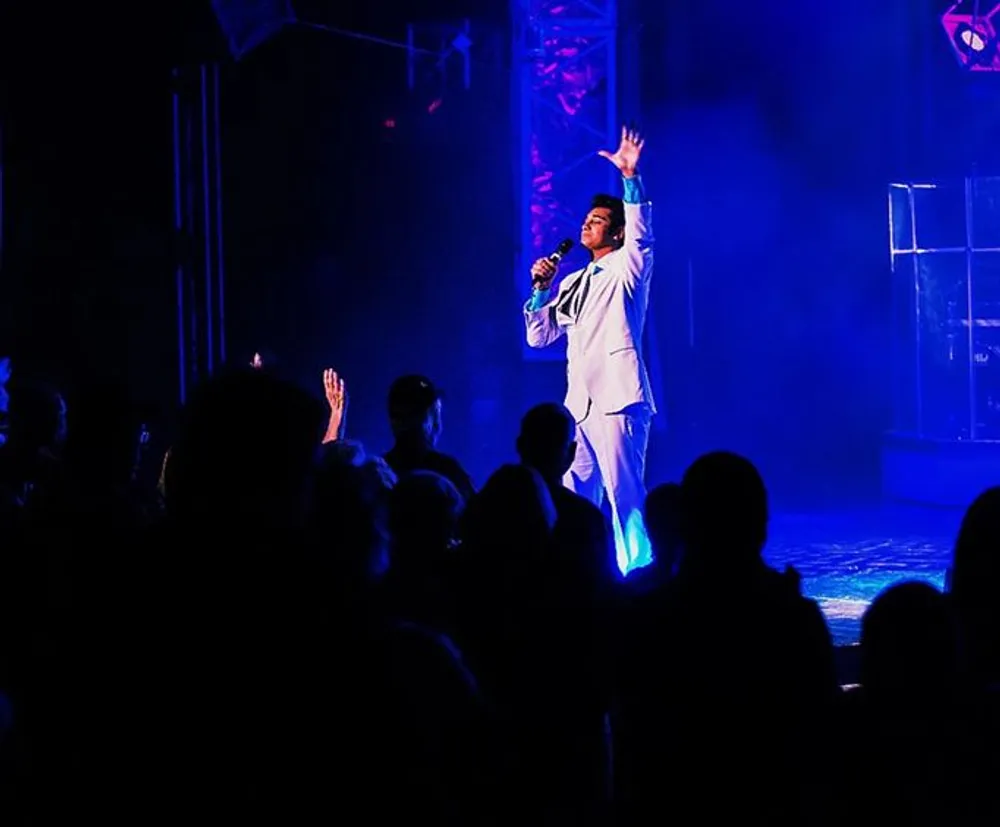 A performer in a white suit is singing on stage with an outstretched arm illuminated by blue stage lights in front of an audience