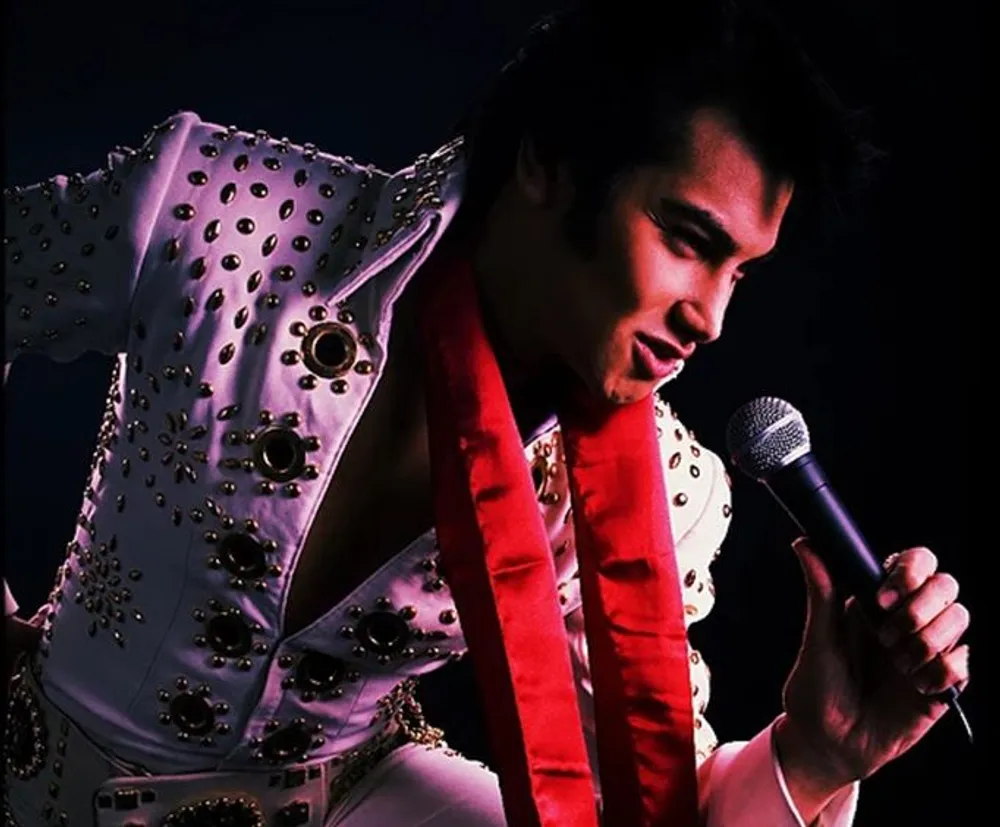 A performer in an elaborate white jumpsuit with rhinestones and a red scarf holds a microphone evoking the style of a classic music era