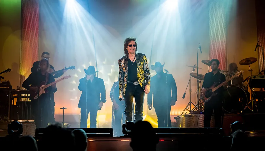 A band performs on stage with a charismatic lead singer in a distinctive jacket standing at the forefront under dramatic stage lighting.