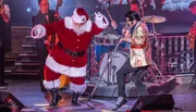 A person dressed as Santa Claus is energetically dancing on stage with a singer in a shiny gold jacket.
