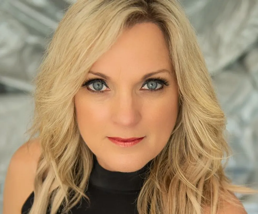 A portrait of a woman with blonde hair blue eyes and wearing a black top with a silvery abstract background