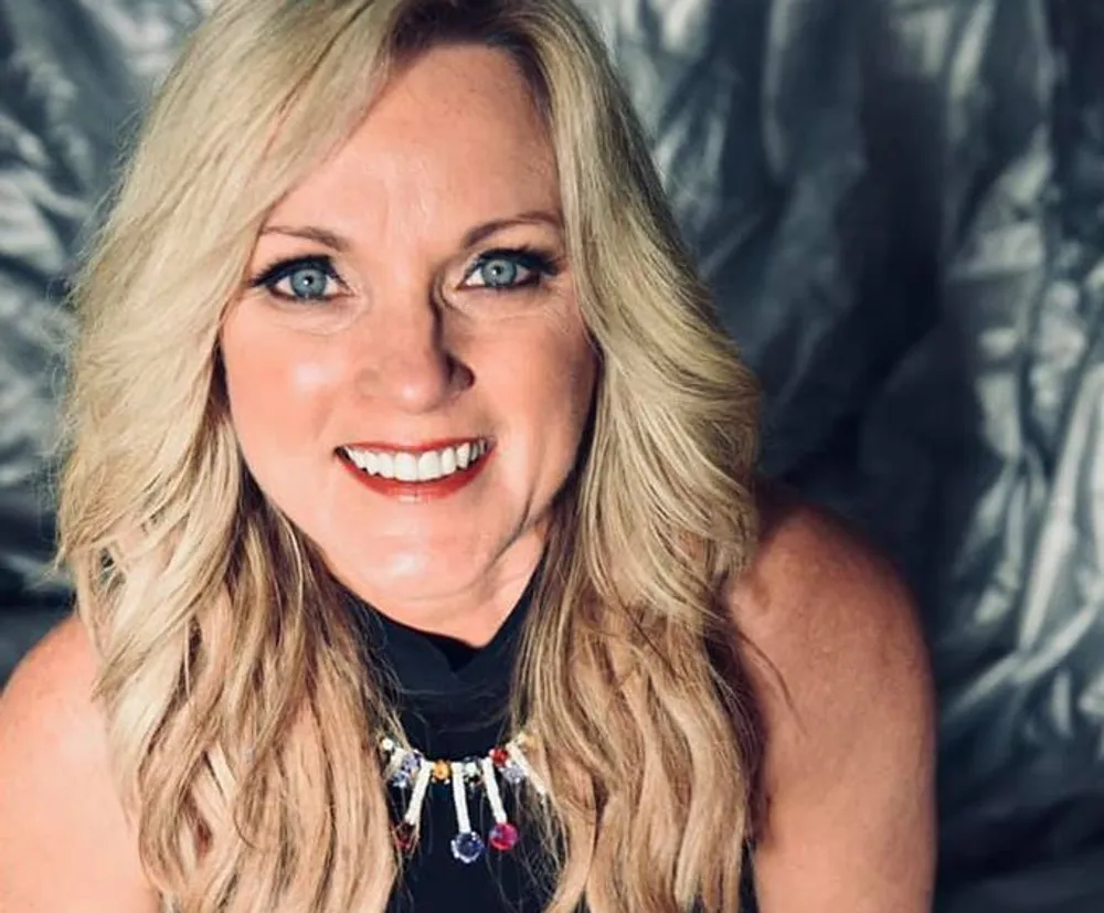 A woman with blonde hair and a bejeweled necklace smiles for a portrait with a silver-toned backdrop