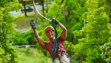 A person wearing a helmet and safety gear is enthusiastically zip-lining through a lush green forest.