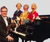 A smiling man in glasses and a tuxedo is seated at a grand piano with a Boston Terrier on the keyboard flanked by three lifelike puppets and a rabbit perched in a top hat