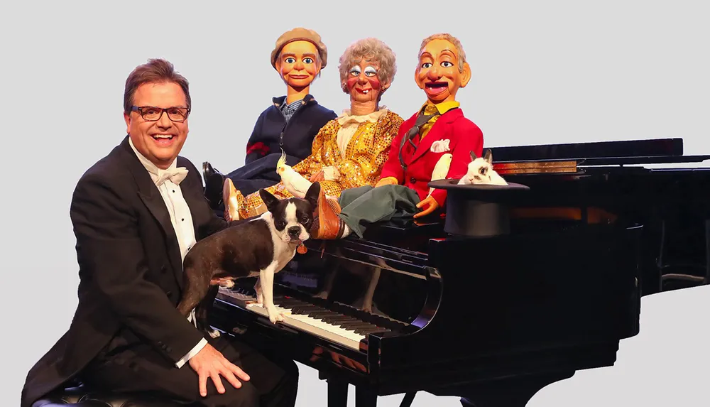 A smiling man in glasses and a tuxedo is seated at a grand piano with a Boston Terrier on the keyboard flanked by three lifelike puppets and a rabbit perched in a top hat