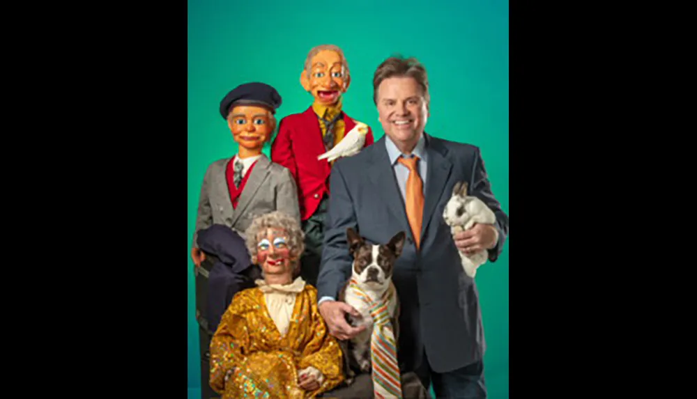 A man in a suit stands holding a dog and a rabbit surrounded by three colorful ventriloquist dummies and a white dove against a green backdrop
