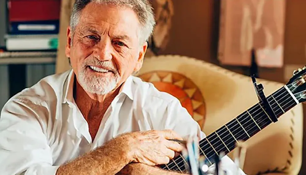 An older man with white hair and a beard is smiling while holding a guitar