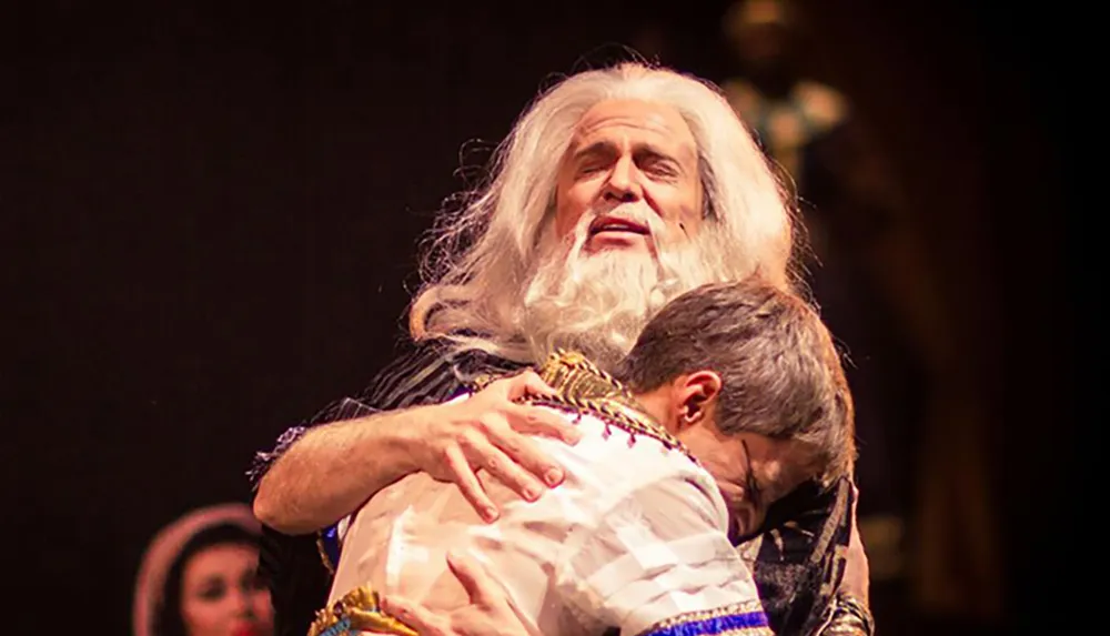 A person with long white hair and a beard is hugging another person on stage in what appears to be a dramatic scene from a theater performance