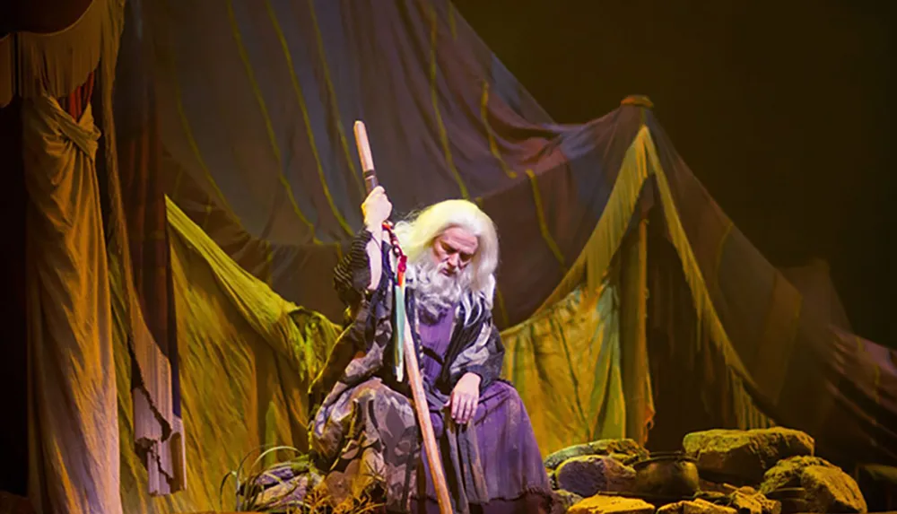 A performer with white hair and beard dressed in a costume with a staff is seated on stage contributing to a scene likely from a theatrical production