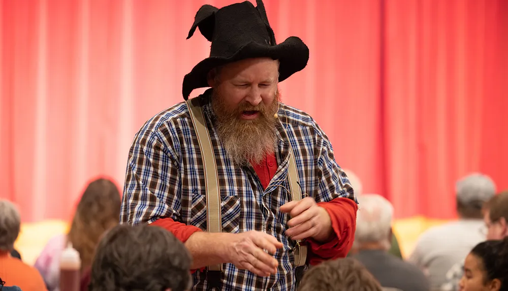 A person dressed in a plaid shirt suspenders and a cowboy-style hat appears to be speaking or performing in front of an audience with a red backdrop
