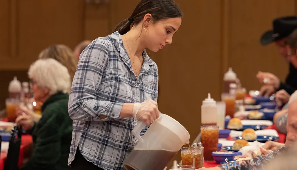 A woman in a plaid shirt is pouring a beverage at a dining event with other attendees eating in the background