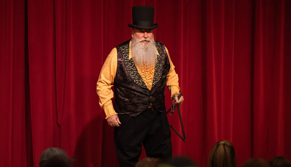 A man with a prominent beard wearing a top hat and traditional showman attire is standing in front of a red curtain holding a whip and appears to be performing in front of an audience