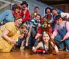 A group of exuberant people in various colorful and characterful costumes pose playfully on a wooden stage likely during a theatrical production or costume party