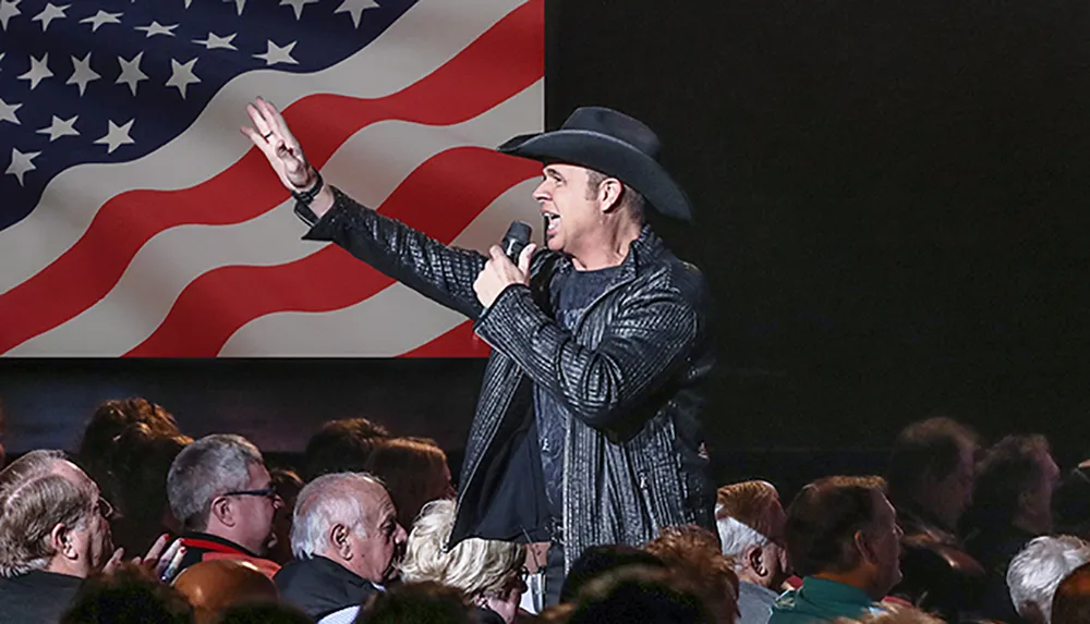 A performer in a cowboy hat is singing into a microphone with an American flag backdrop