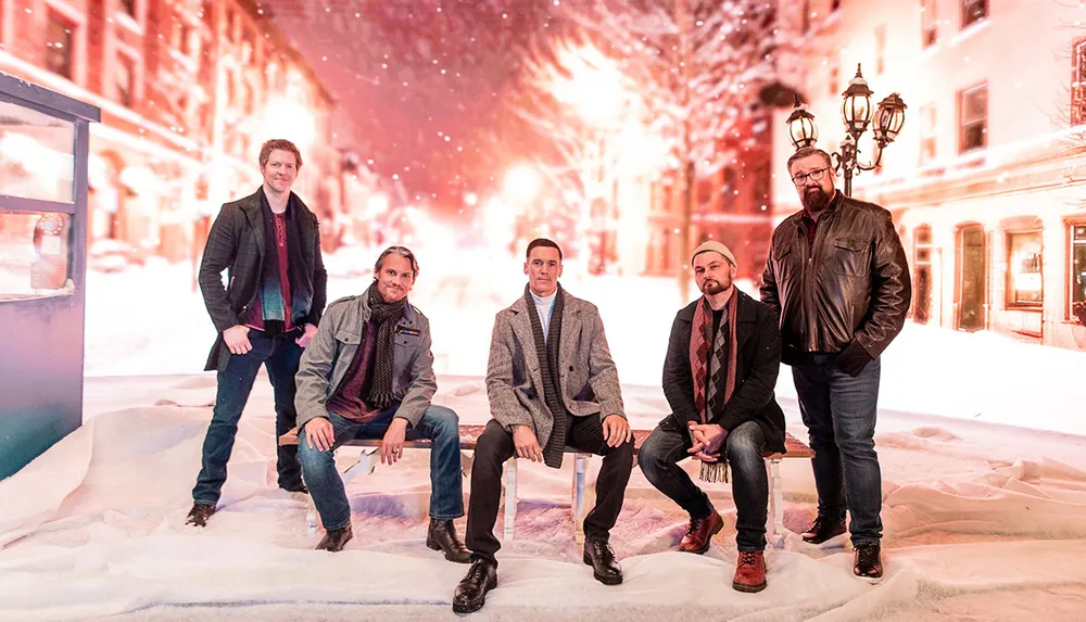 Five men wearing winter clothing are posing in front of a snowy illuminated street scene