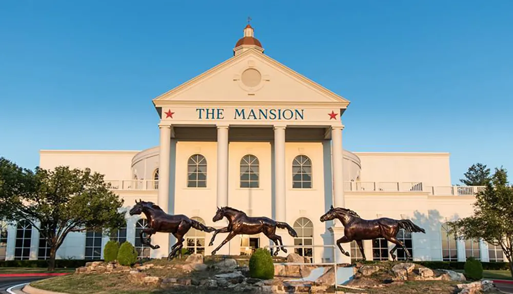 The image shows a grand white building with the words THE MANSION at the front flanked by two star emblems and featuring a fountain with bronze horse sculptures in the foreground