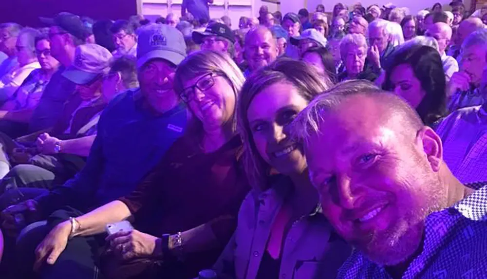 A group of people takes a smiling selfie together in a crowded auditorium or event venue