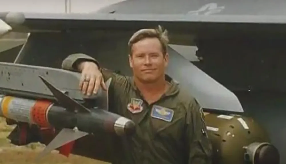 A person in a military flight suit is leaning on an aircraft with a missile attached to it smiling at the camera