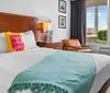 The image shows a neatly arranged hotel room with a bed featuring a yellow pillow that reads Today is a good day a turquoise throw an armchair a bedside table with a lamp and a window revealing a view outside