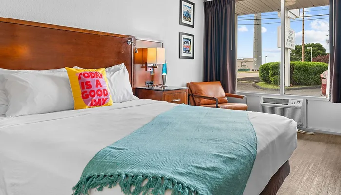 The image shows a neatly arranged hotel room with a bed featuring a yellow pillow that reads Today is a good day a turquoise throw an armchair a bedside table with a lamp and a window revealing a view outside