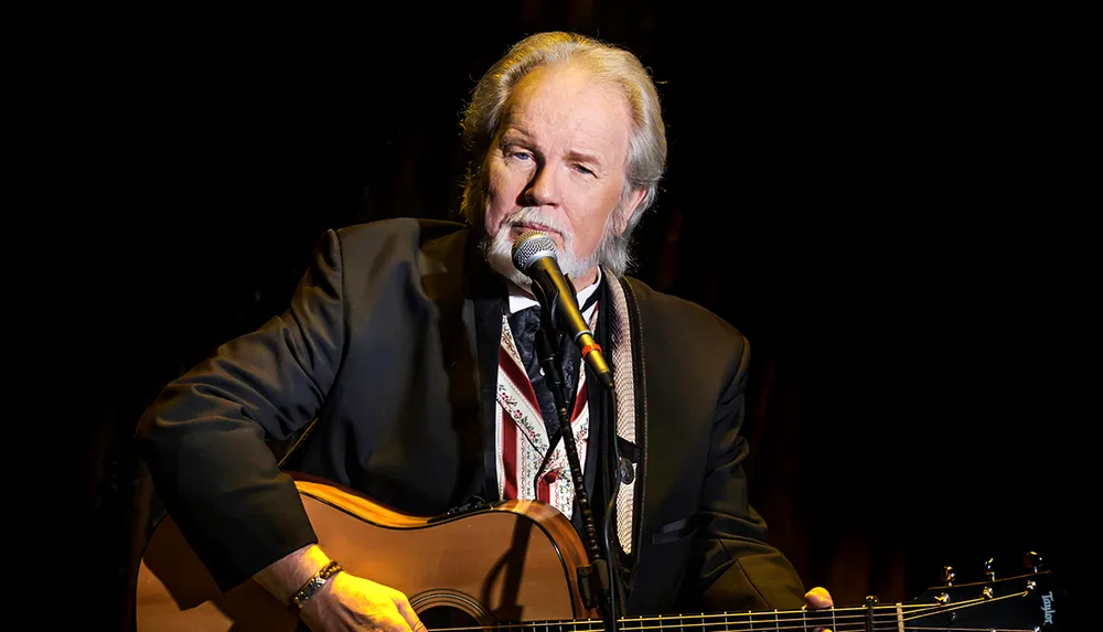 A man is playing an acoustic guitar and singing into a microphone on stage wearing a suit