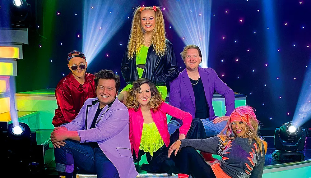 Six people dressed in colorful retro-inspired outfits are posing together on a vibrant stage set