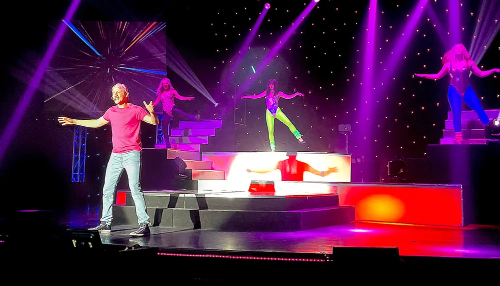 A man in a red shirt is energetically performing on a brightly lit stage with three women in colorful outfits dancing in the background