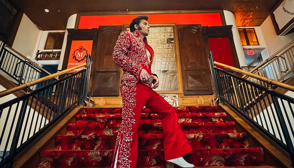 A person stands confidently on a red-carpeted staircase wearing a detailed red suit with ornate embellishments