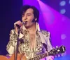 A performer dressed in a sequined outfit is singing into a microphone on stage illuminated by colorful lights with a guitar in their hand