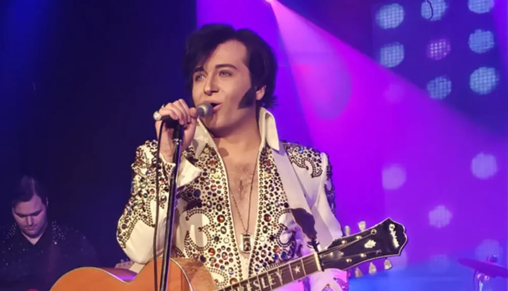A performer dressed in a sequined outfit is singing into a microphone on stage illuminated by colorful lights with a guitar in their hand