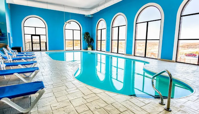 An indoor swimming pool with a vibrant blue color scheme is surrounded by arched windows and lounge chairs
