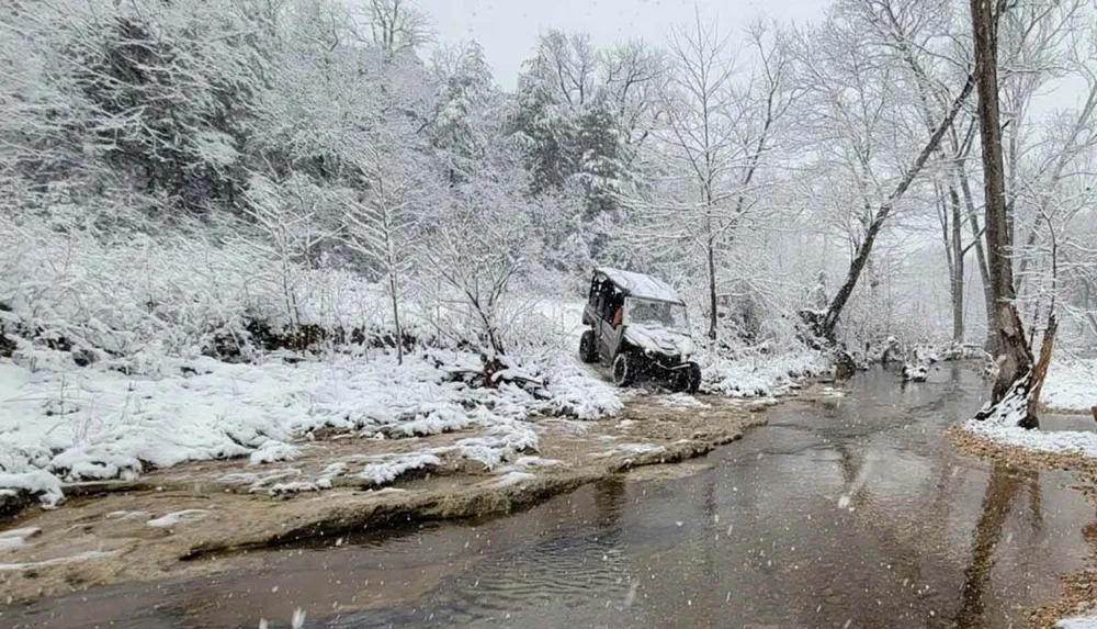 A snowy landscape with a four-wheeled vehicle parked at the side of a winding water-covered road surrounded by snow-covered trees