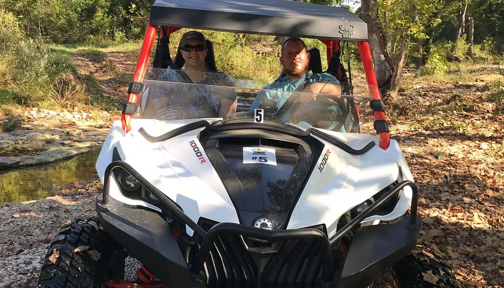 Two individuals are sitting in a side-by-side off-road utility vehicle on a trail in a wooded area