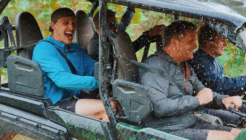 Three people are enjoying a rain-soaked ride in an off-road vehicle with the driver and one passenger smiling widely as they are splashed with water
